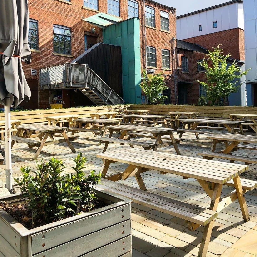 Outdoor seating area with wooden picnic tables at The Cross Keys, surrounded by brick buildings and greenery on a sunny day.
