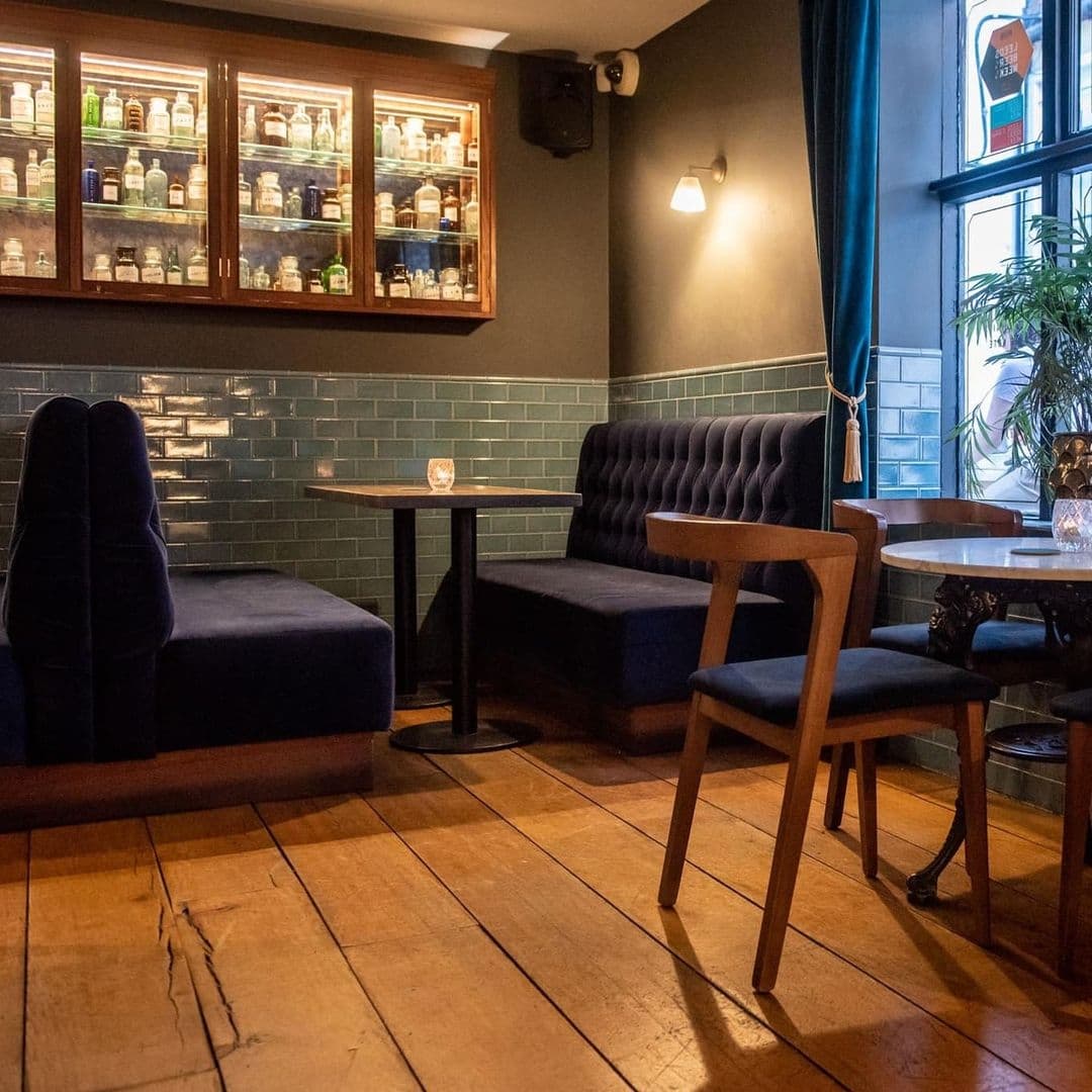 Interior of The Turk’s Head featuring a cozy seating area with blue upholstered benches and chairs, wooden tables, and a well-stocked bar cabinet against a tiled wall.
