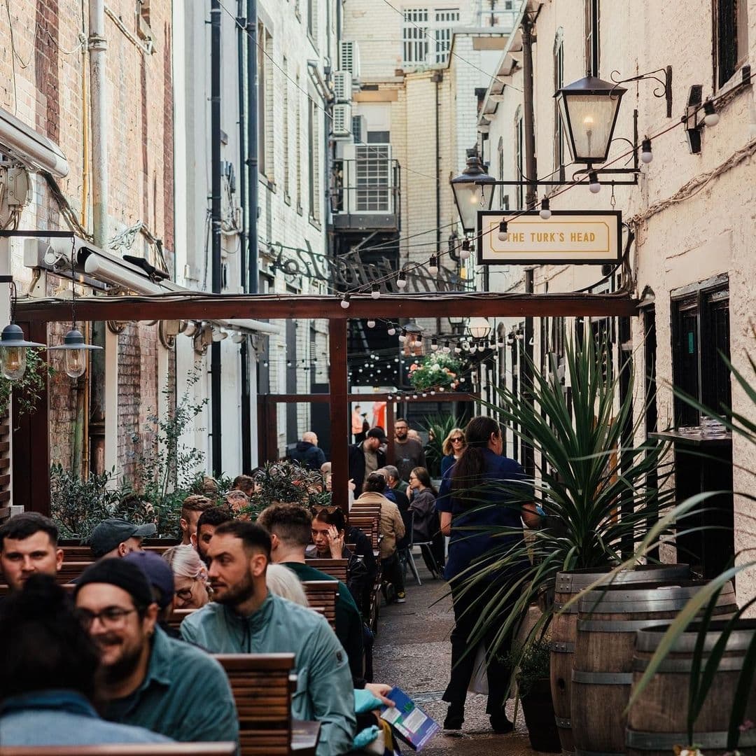 Outdoor seating area at The Turk's Head with people dining, surrounded by brick buildings, green plants, and string lights.