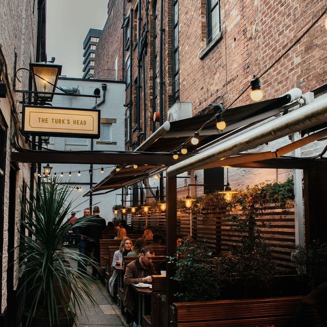 Outdoor seating area of The Turk's Head with patrons dining under string lights, surrounded by brick buildings.