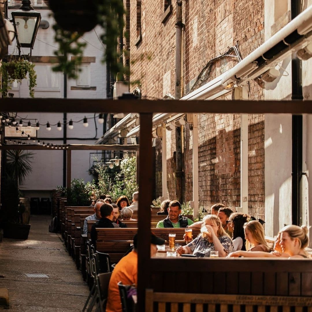 Outdoor seating at The Turk's Head with people enjoying drinks in a cozy alleyway surrounded by brick buildings and hanging lights.