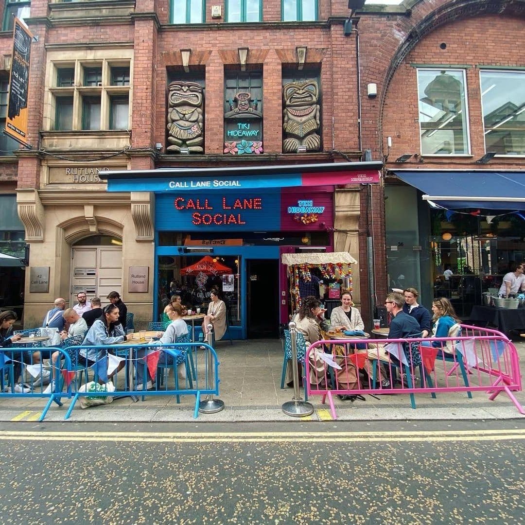 Exterior view of Tiki Hideaway with patrons dining at colorful tables on the sidewalk, adjacent to the Call Lane Social entrance.
