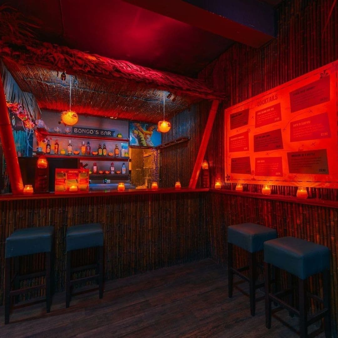 Interior of Tiki Hideaway with bamboo bar, red ambient lighting, tiki-themed decorations, and cocktail menu on wall.