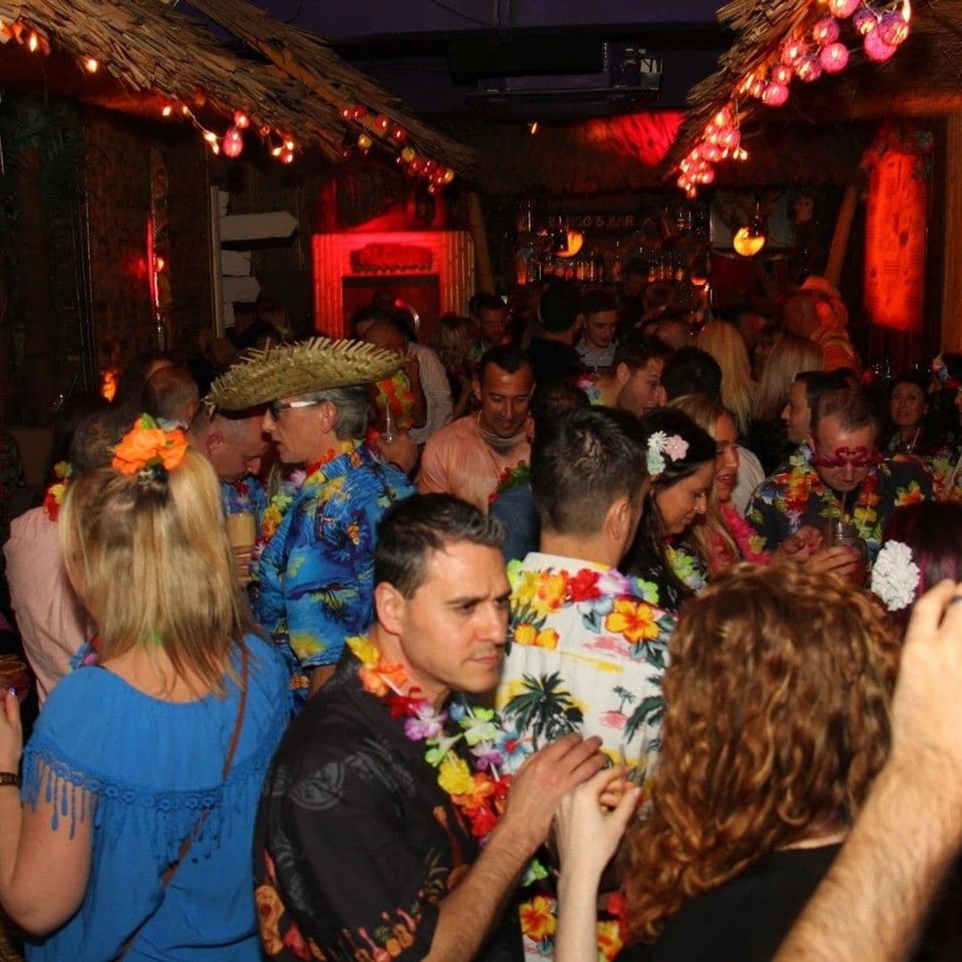 Crowd of people enjoying a lively party at Tiki Hideaway, wearing colorful Hawaiian shirts and floral leis, with tropical-themed decor in the background.