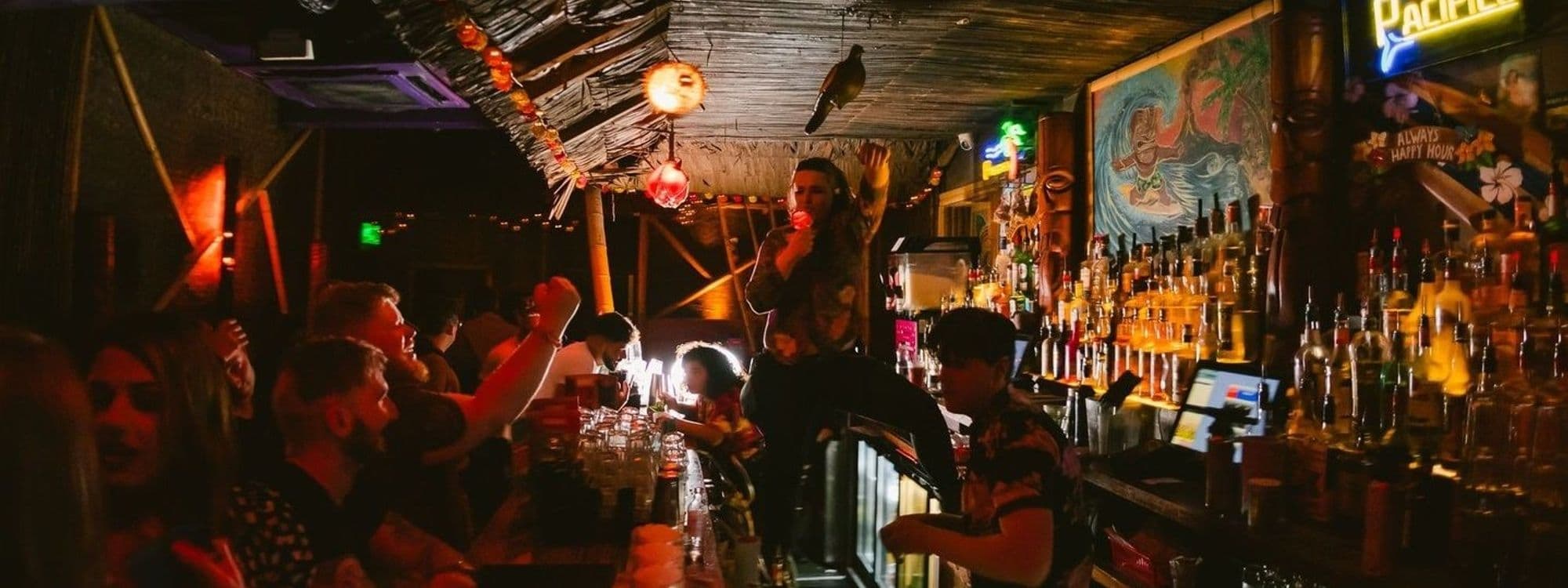 Vibrant nightlife scene at Tiki Hideaway with patrons enjoying drinks and socializing at the bar adorned with tropical decor and dim, warm lighting.