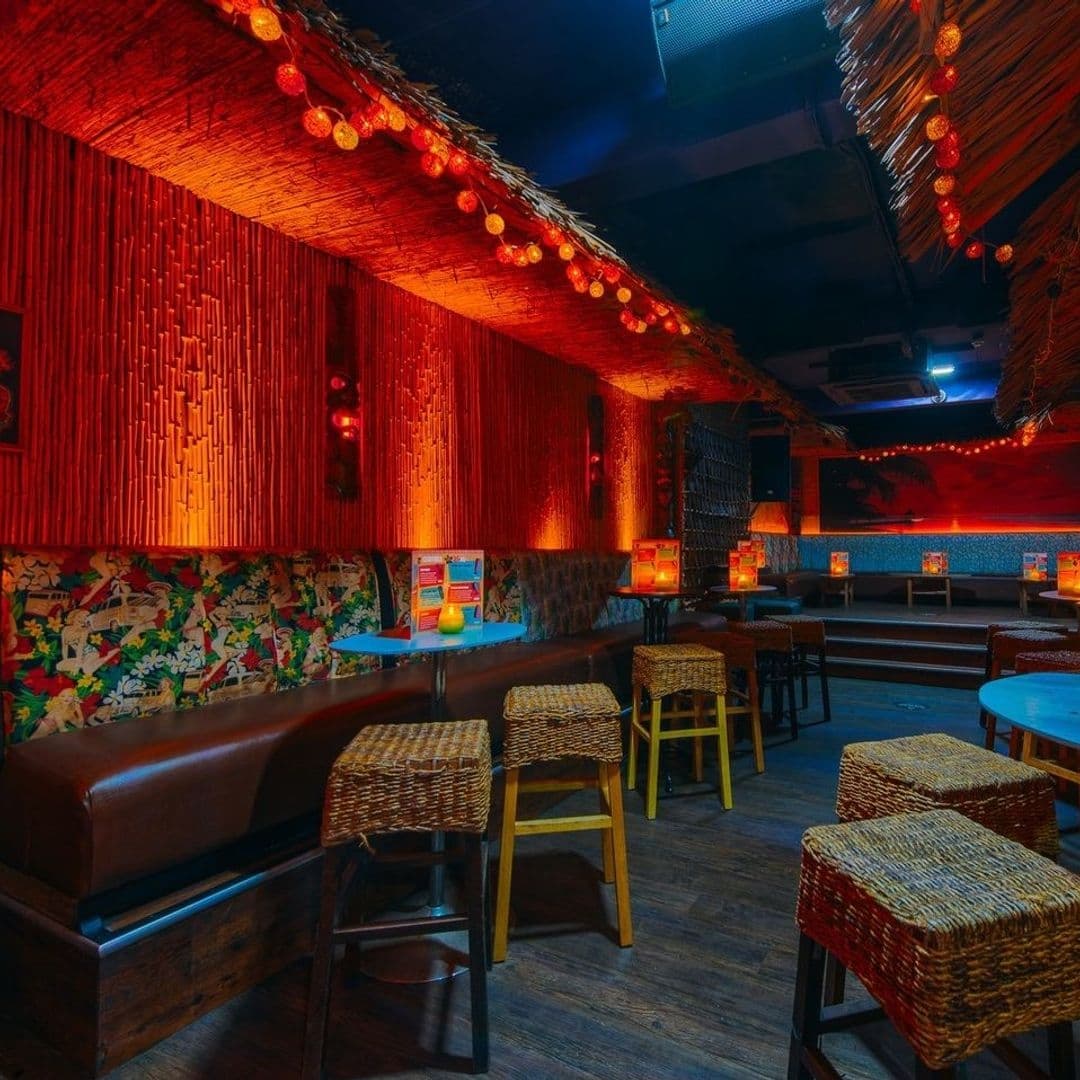 Interior of Tiki Hideaway with tropical-themed decor, featuring bamboo-lined walls, thatched roof accents, colorful cushioned benches, wicker bar stools, and warm ambient lighting.