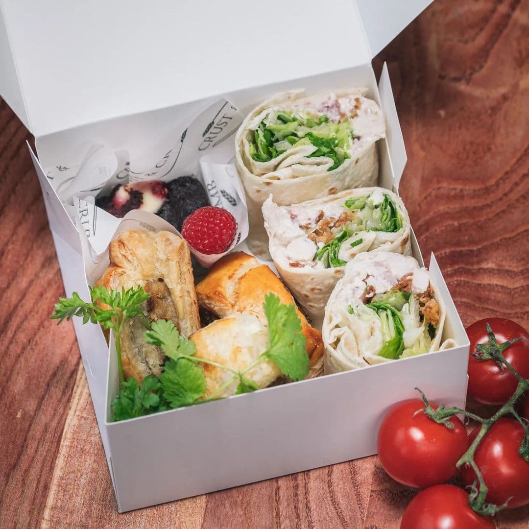 Open white takeout box from Crust & Crumb containing an assortment of fresh wraps, pastries, and berries on a wooden surface with ripe tomatoes alongside.