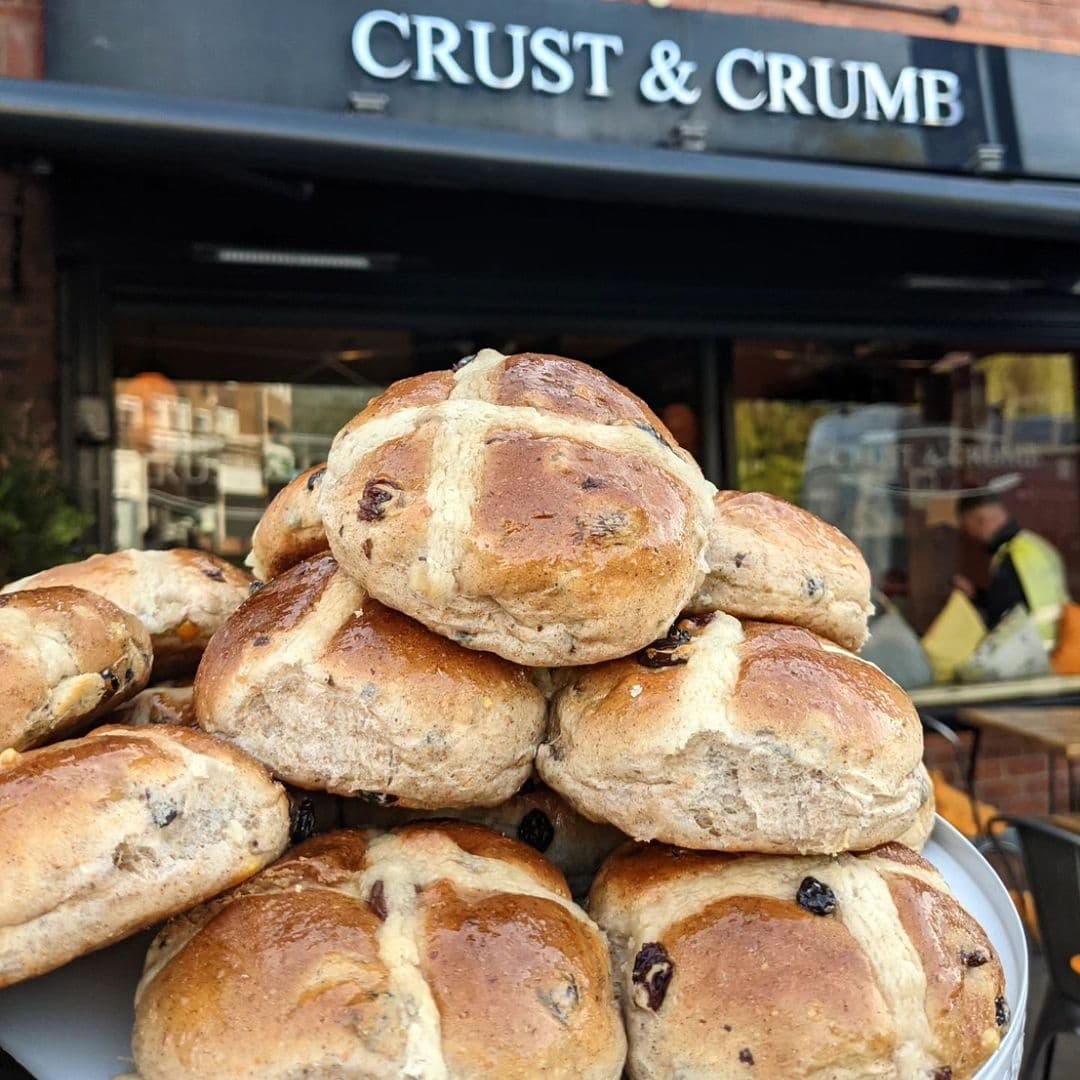 Freshly baked hot cross buns piled on a plate in front of the Crust & Crumb bakery, with the shop's sign visible in the background.