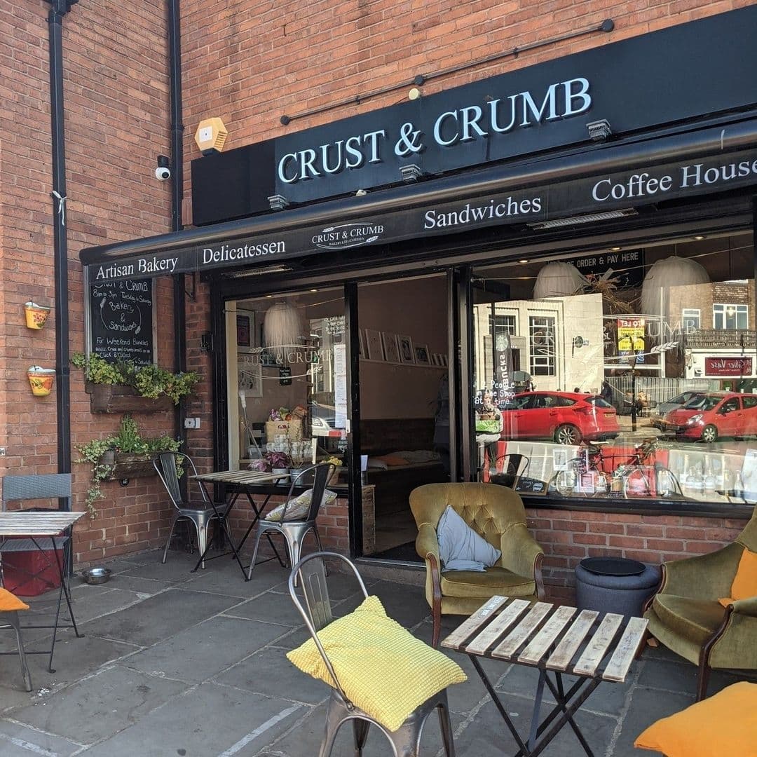 Exterior view of Crust & Crumb artisan bakery and coffee house with outdoor seating and open entrance on a sunny day.