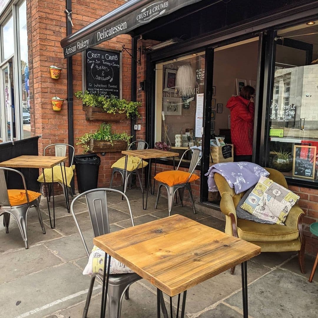 Outdoor seating area of Crust & Crumb bakery with wooden tables, metal chairs, and a cozy armchair, in front of the shop with open door and a person inside. Decorative plants and a blackboard with menu items are visible.