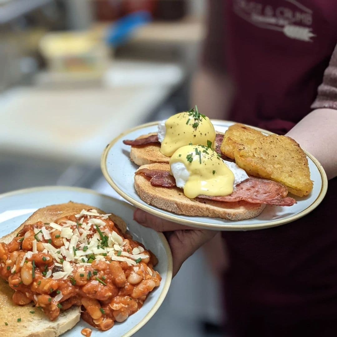 "Savory breakfast dishes served at Crust & Crumb, featuring eggs benedict with bacon and hash browns on one plate, and beans on toast sprinkled with cheese on another, held by a staff member in a maroon apron."