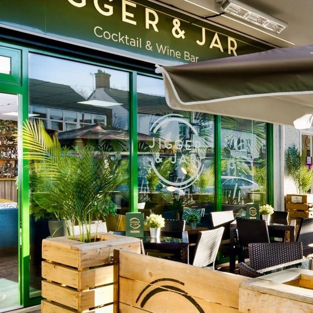 Outdoor seating area at Jigger & Jar Cocktail & Wine Bar with rattan chairs, wooden tables, and lush green plants.
