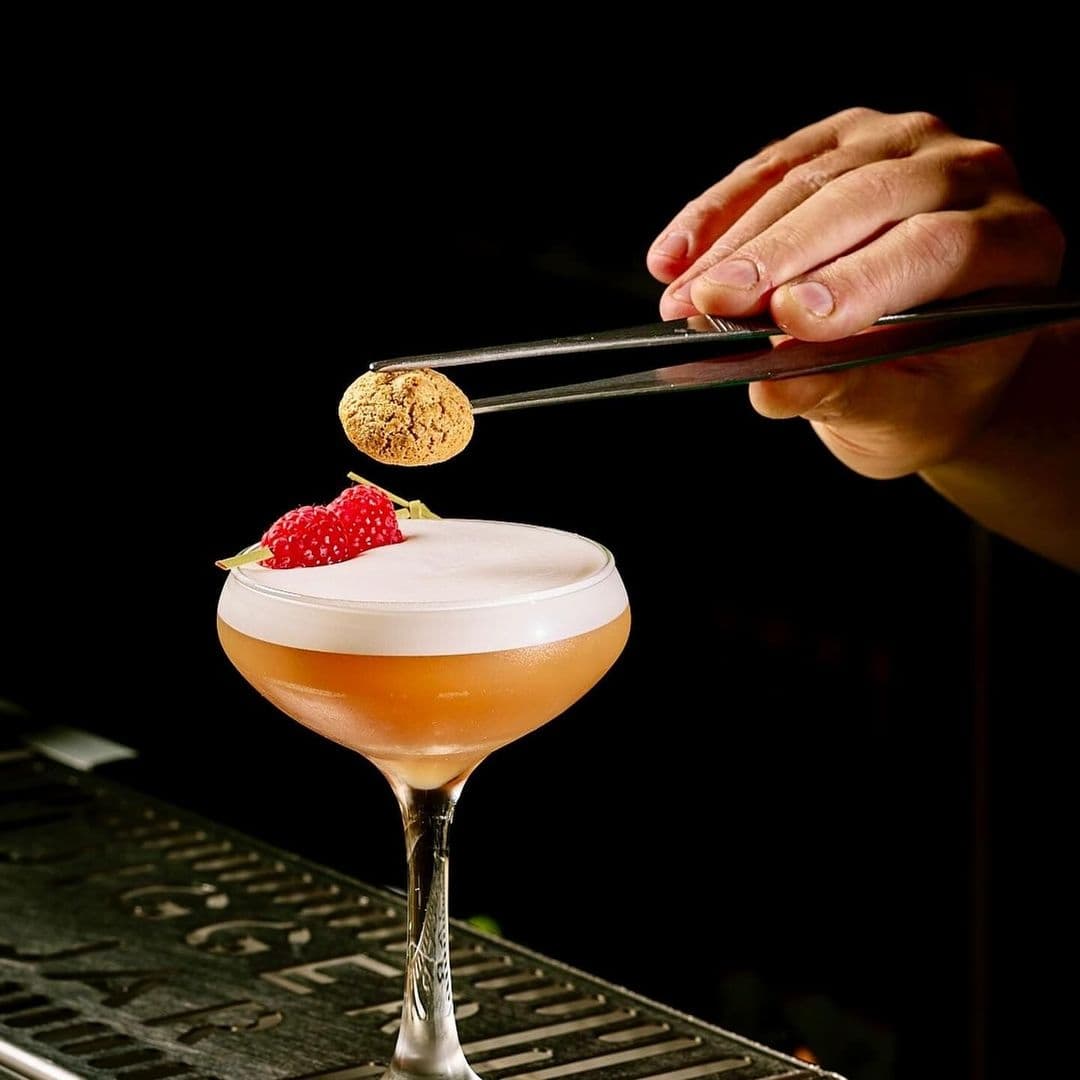 Alt text: "Hand using tweezers to garnish a frothy cocktail with a cookie and raspberry at Jigger & Jar."