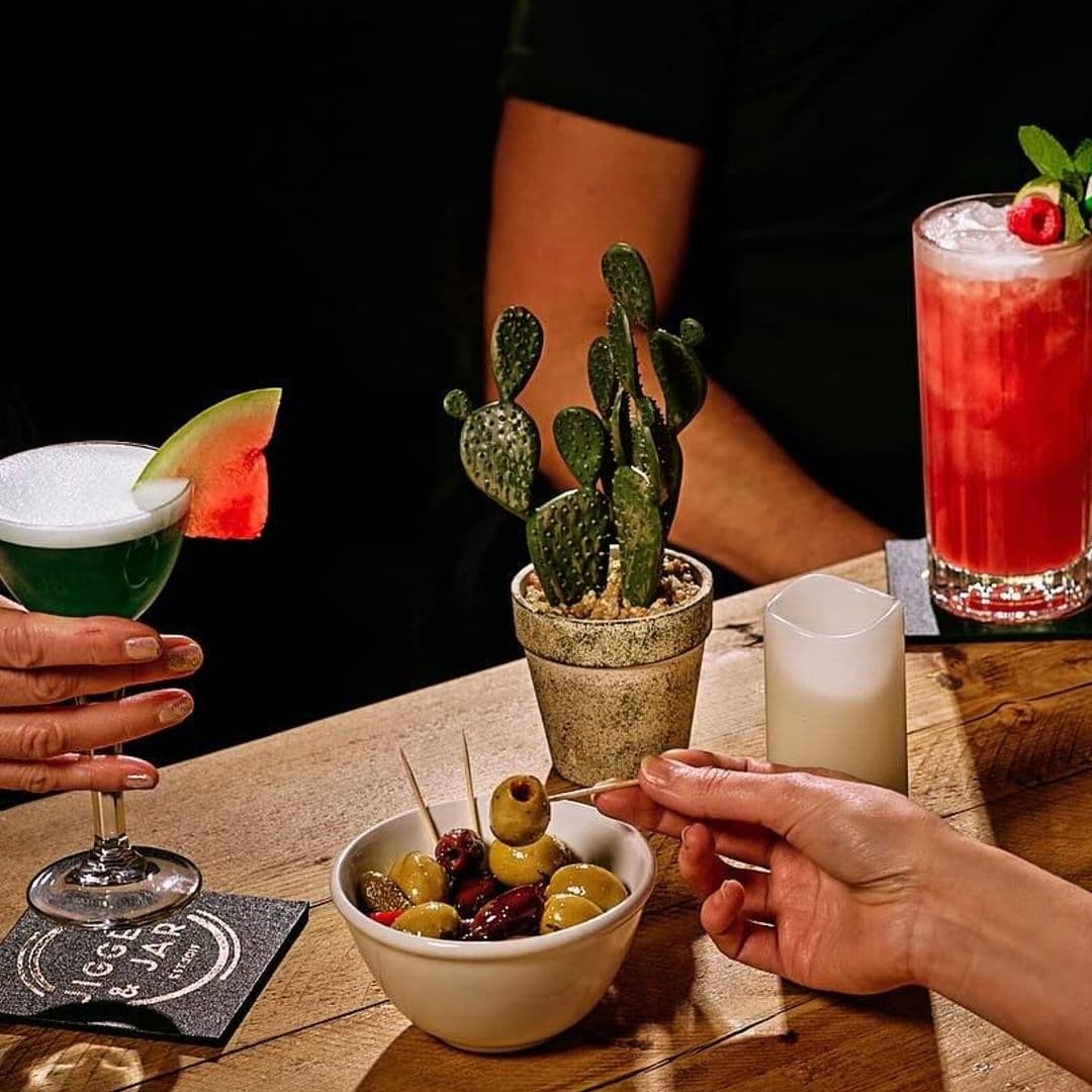 "Patrons enjoying vibrant cocktails and a bowl of mixed olives at Jigger & Jar, with a potted cactus centerpiece on a wooden table."