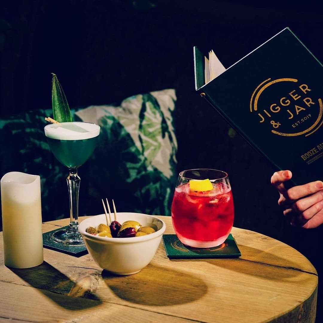 "Inviting cocktail setup at Jigger & Jar featuring a vibrant red drink with lemon garnish, a green cocktail with a decorative leaf, a bowl of olives, and a menu held by a person in the background."