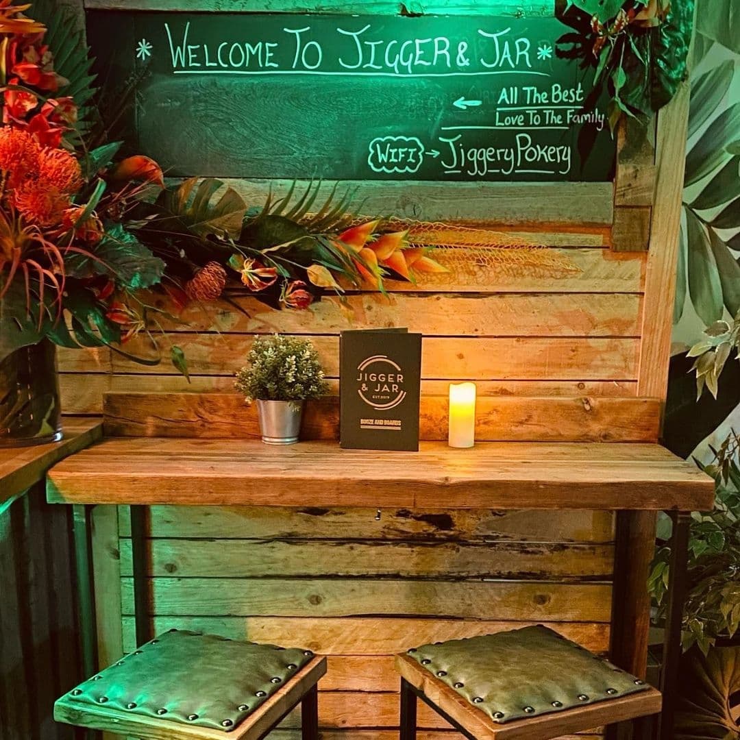 Cozy corner at Jigger & Jar with wooden bench, metal stools, a lit candle, and a welcoming chalkboard sign surrounded by warm lighting and greenery.