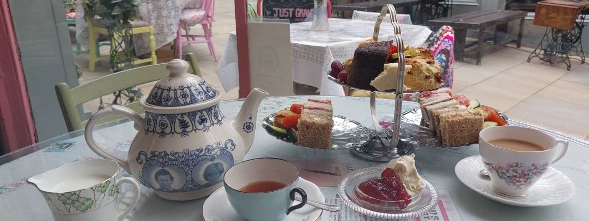 Traditional afternoon tea setup at Just Grand! Vintage Tearoom, featuring an ornate blue and white teapot, matching cup with tea, a selection of finger sandwiches, scones, and cakes on a tiered stand, with a heart-shaped jelly and cream dessert in the foreground.