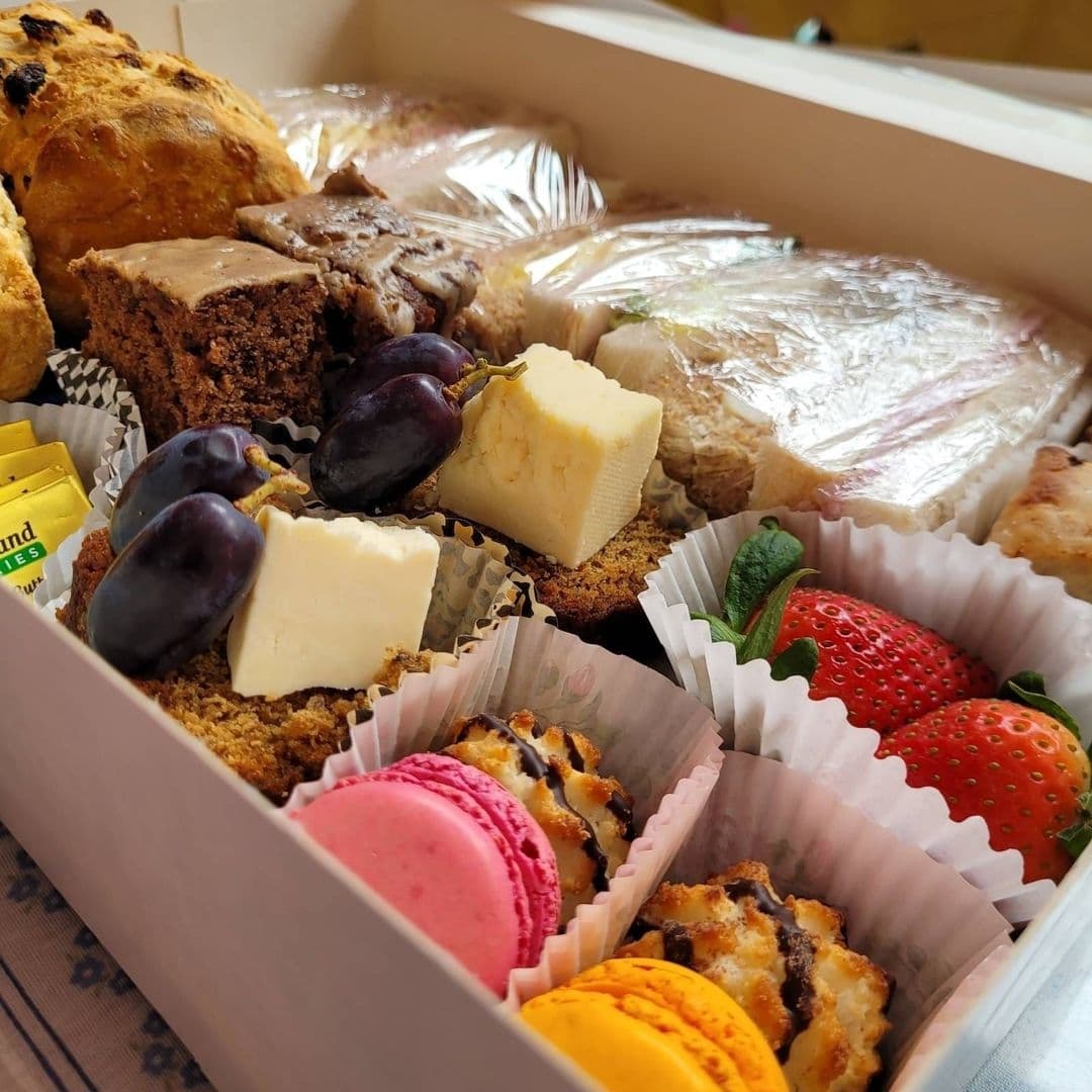 Assorted pastries and treats from Just Grand! Vintage Tearoom, including scones, brownies, macarons, cheese cubes, grapes, and strawberries, presented in a box.