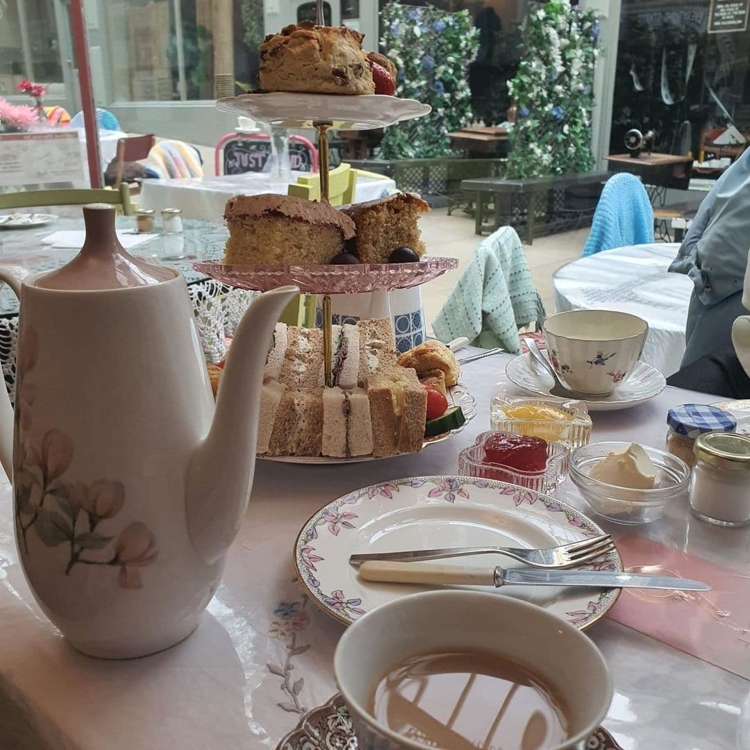 Afternoon tea setup at Just Grand! Vintage Tearoom with a three-tiered cake stand filled with scones, cakes, and sandwiches, a floral porcelain teapot, cups, and condiments on a lace tablecloth, with patrons and greenery in the background.