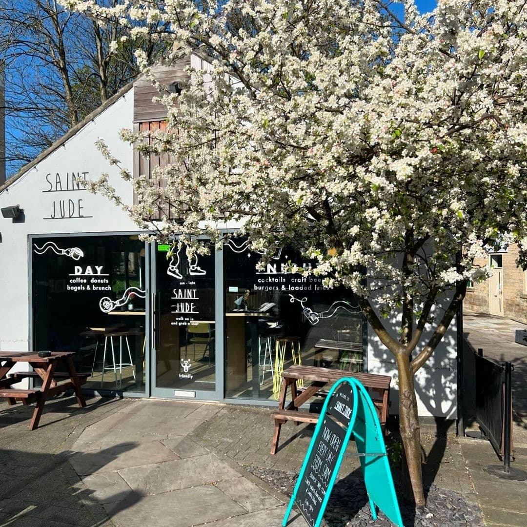 Exterior view of Saint Jude cafe with blooming white flowers on a tree in the foreground, wooden benches outside, and a blue sidewalk sign featuring menu items.
