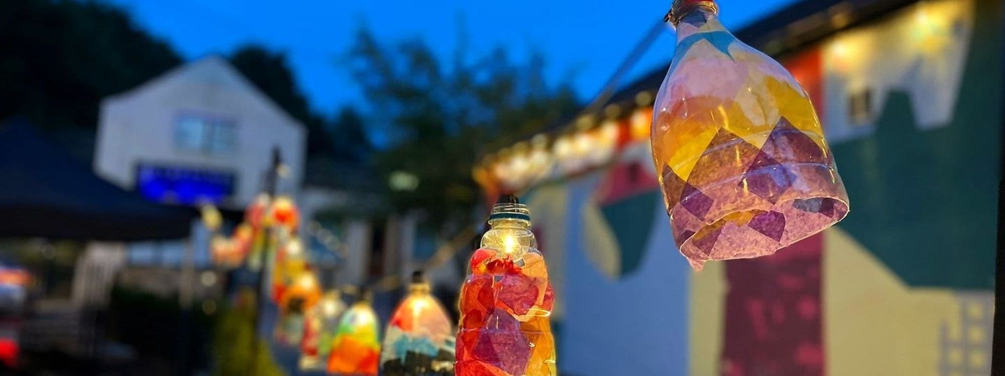 Colorful hanging lanterns at a festive outdoor event in Saint Jude, with a blurred background of buildings and trees at dusk.