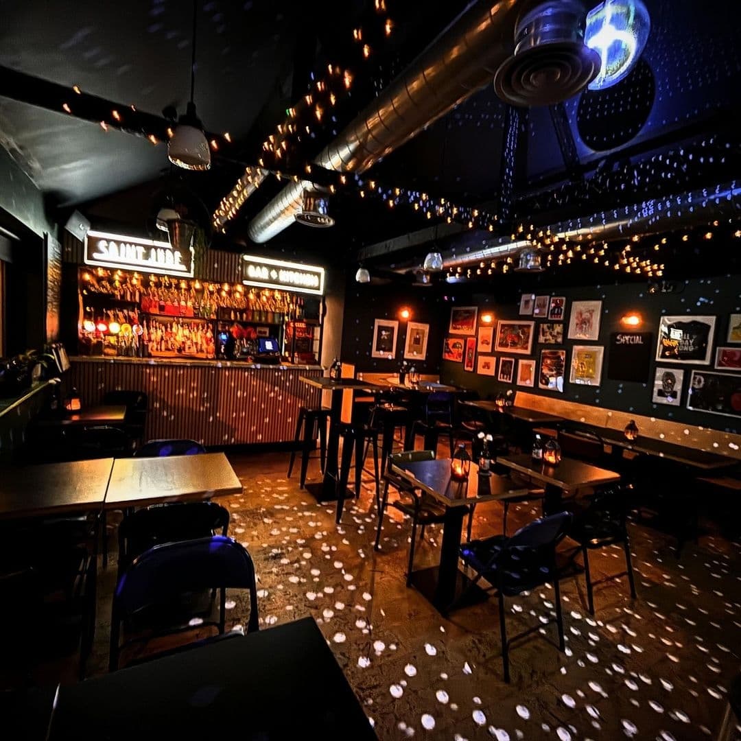 Interior of Saint Jude bar with ambient lighting, featuring a well-stocked bar counter, framed pictures on the wall, and scattered seating tables under a ceiling adorned with fairy lights.