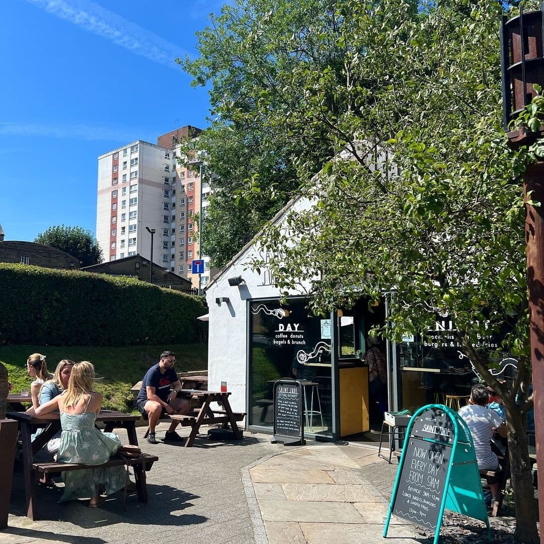 Outdoor seating at a cozy café in Saint Jude with patrons enjoying the sunny weather, surrounded by greenery and a high-rise building in the background.
