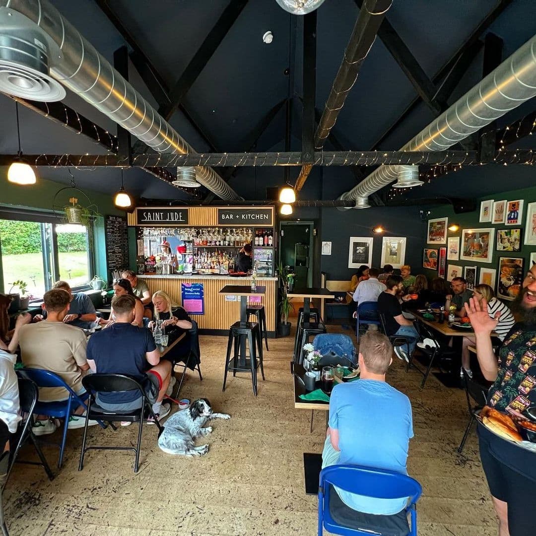 Interior of Saint Jude bar and kitchen with patrons dining and socializing, featuring a well-stocked bar and contemporary decor. A dog is lying on the floor, adding to the casual, pet-friendly atmosphere.