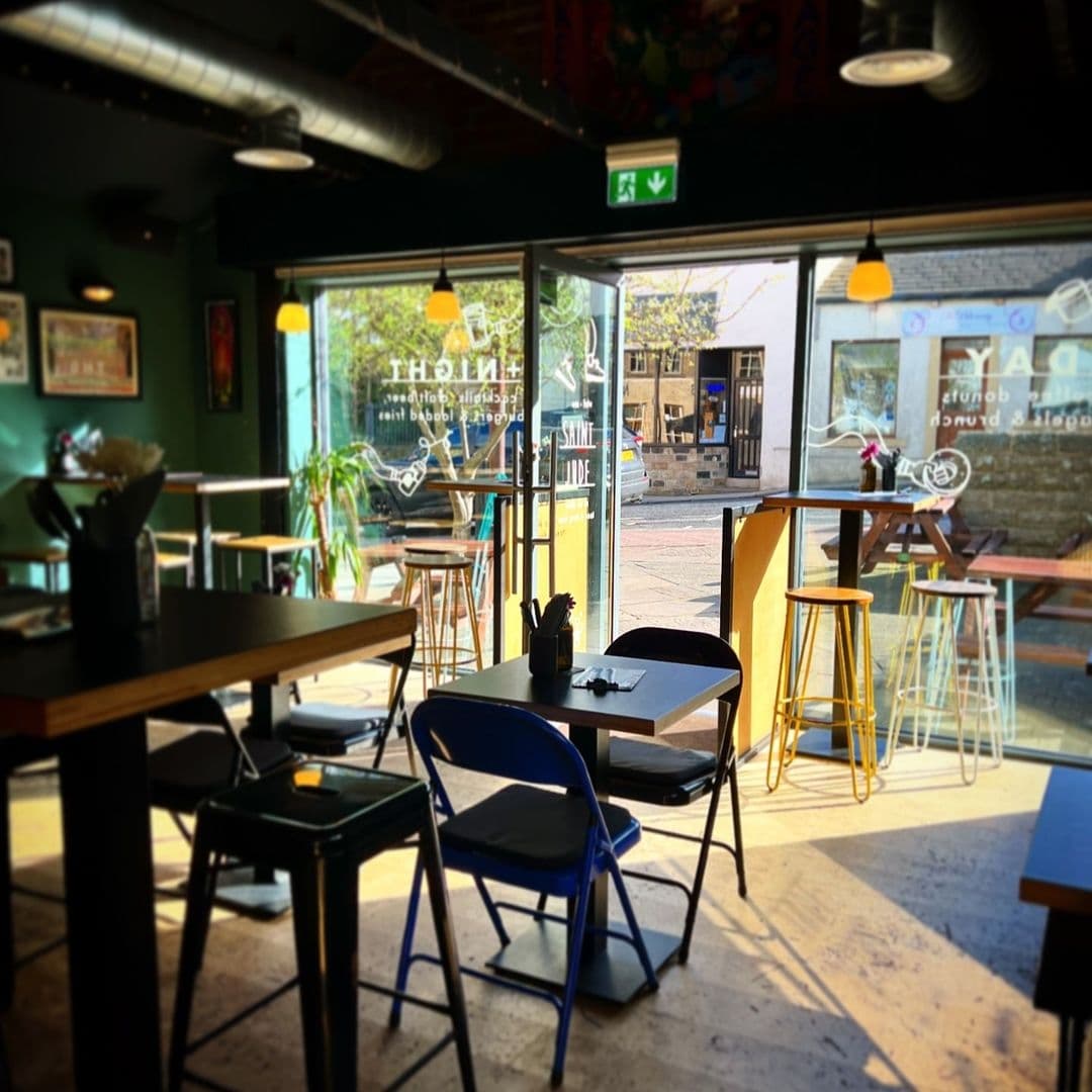 Interior of Saint Jude cafe with wooden tables, blue and black chairs, and yellow stools near the entrance, which is adorned with large windows offering a view of the street outside.