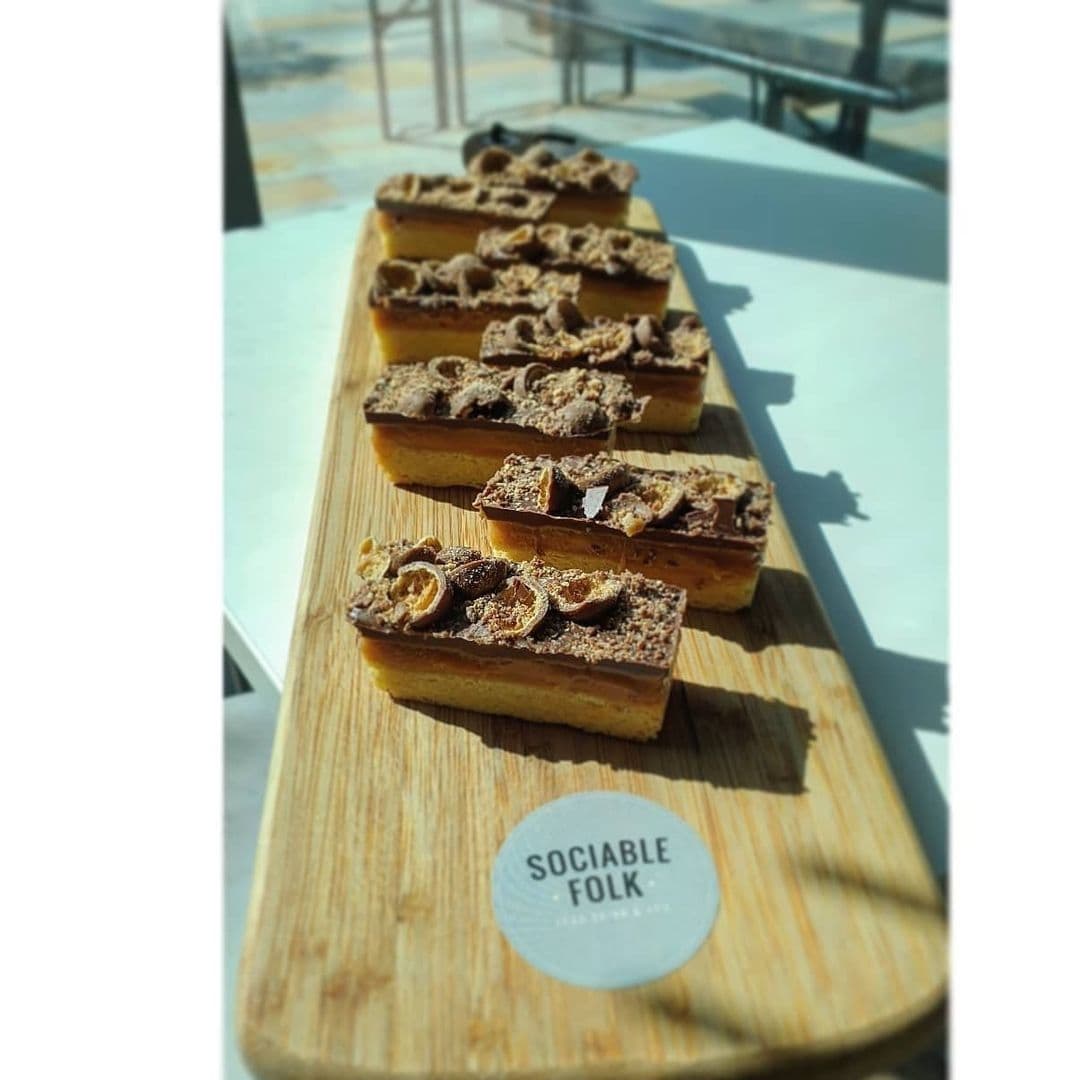 A wooden board with a row of freshly made caramel shortbread slices topped with chocolate pieces, displayed at Sociable Folk cafe.