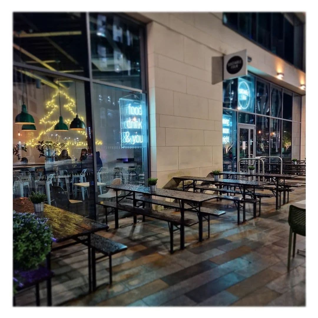 Outdoor seating area at Sociable Folk with empty tables and chairs on a patterned pavement, illuminated by warm interior lighting and decorative string lights visible through the cafe's large glass windows.
