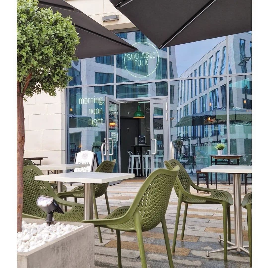 Outdoor seating area at Sociable Folk with green chairs and tables under an umbrella, a potted tree, and a view of the modern glass building facade in the background.
