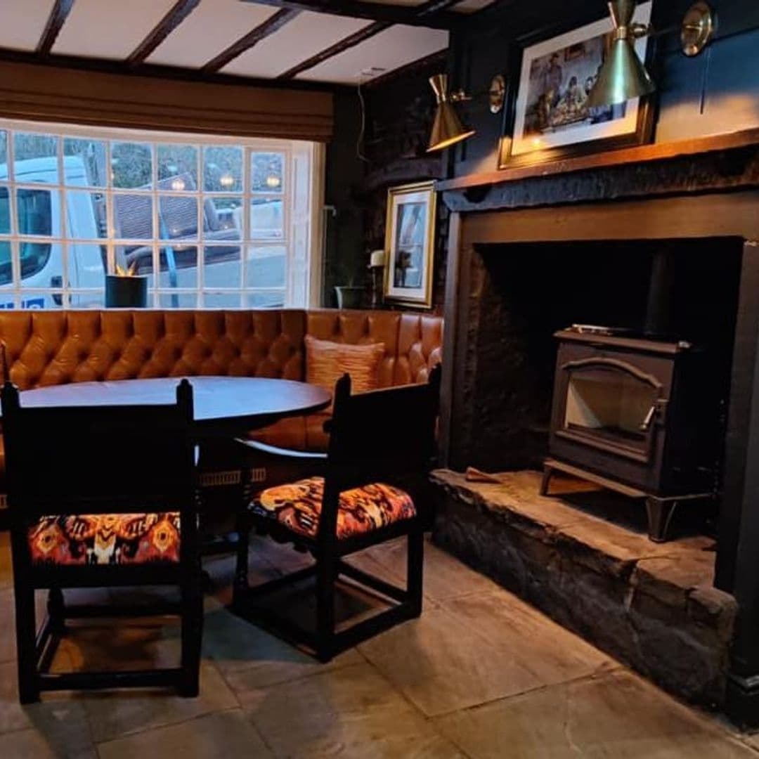 Cozy interior of the Bankhouse Inn featuring a tufted leather booth, wooden chairs with patterned cushions, a wood stove fireplace, and framed pictures on dark walls, creating a warm and inviting atmosphere.