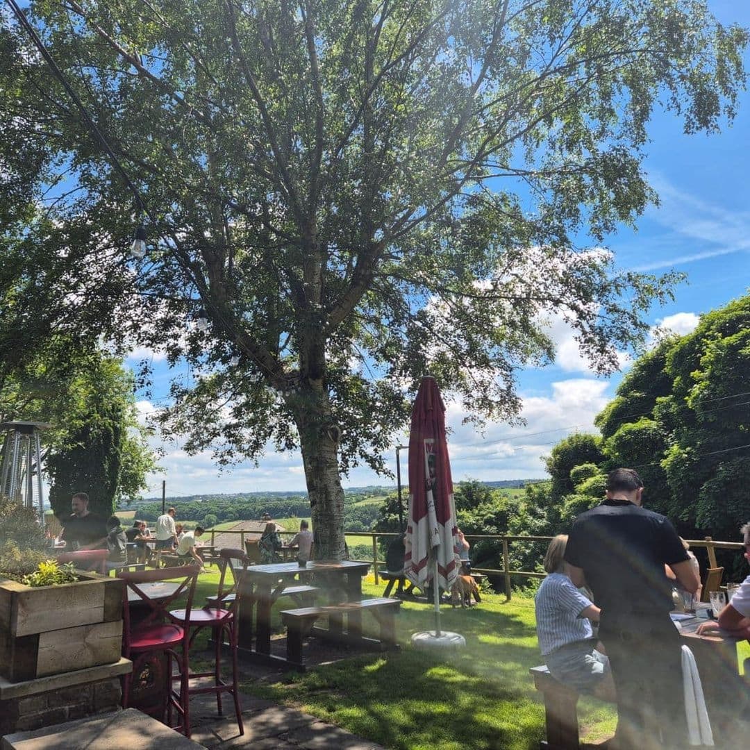Outdoor seating area at Bankhouse Inn with patrons enjoying the sunny weather under the shade of a large tree, overlooking scenic countryside views.