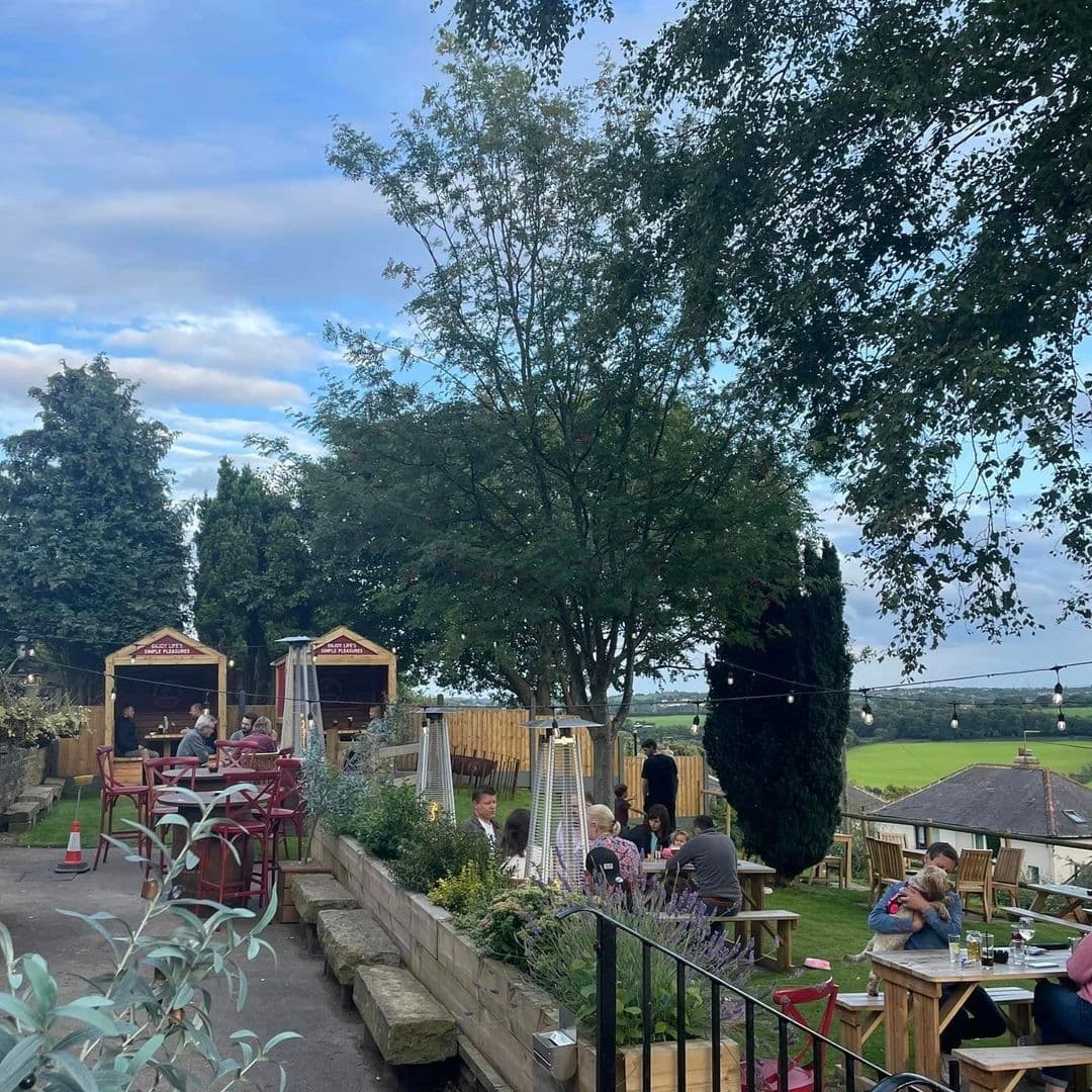 Outdoor seating area at Bankhouse Inn with guests enjoying meals, wooden huts, string lights, and a view of greenery in the background.