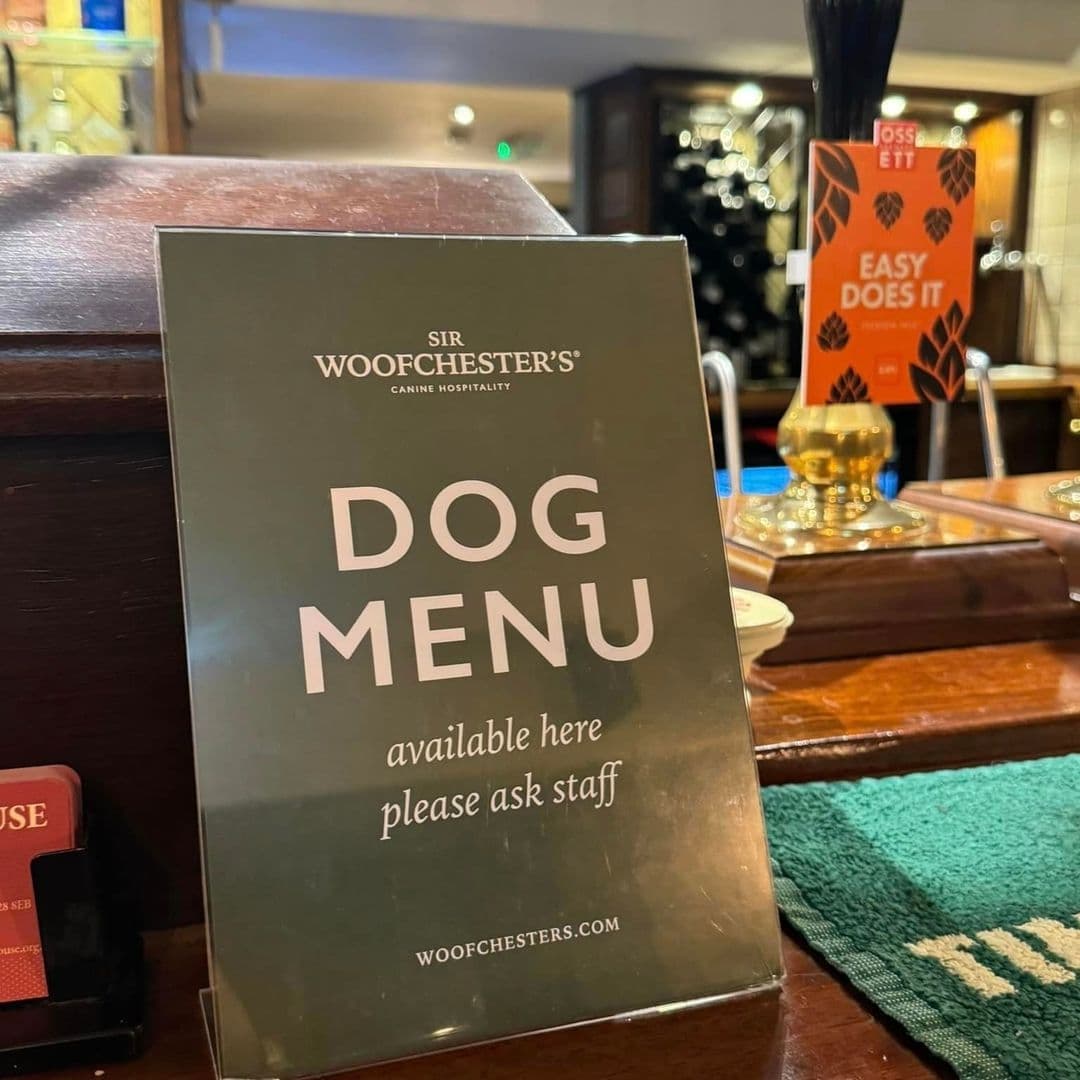 Dog menu sign on a wooden bar counter at Bankhouse Inn, indicating pet-friendly services with a beer tap in the background.