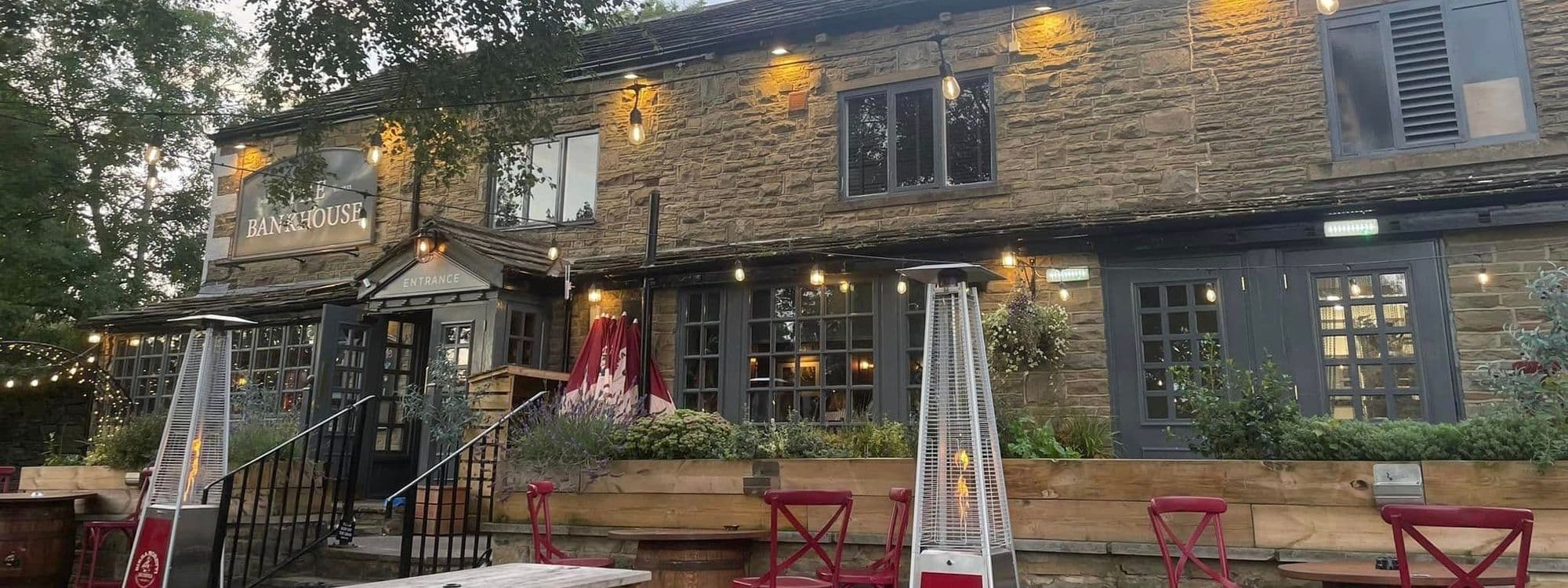 Exterior view of Bankhouse Inn with illuminated signage, stone facade, and outdoor seating area featuring red stools, wooden tables, and patio heaters.
