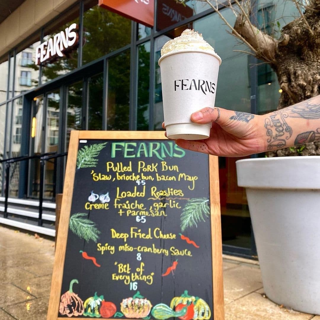 Hand holding a coffee cup with "FEARNS" logo in front of Fearns Dock restaurant, with a menu board showcasing food items like Pulled Pork Bun and Loaded Roasties.