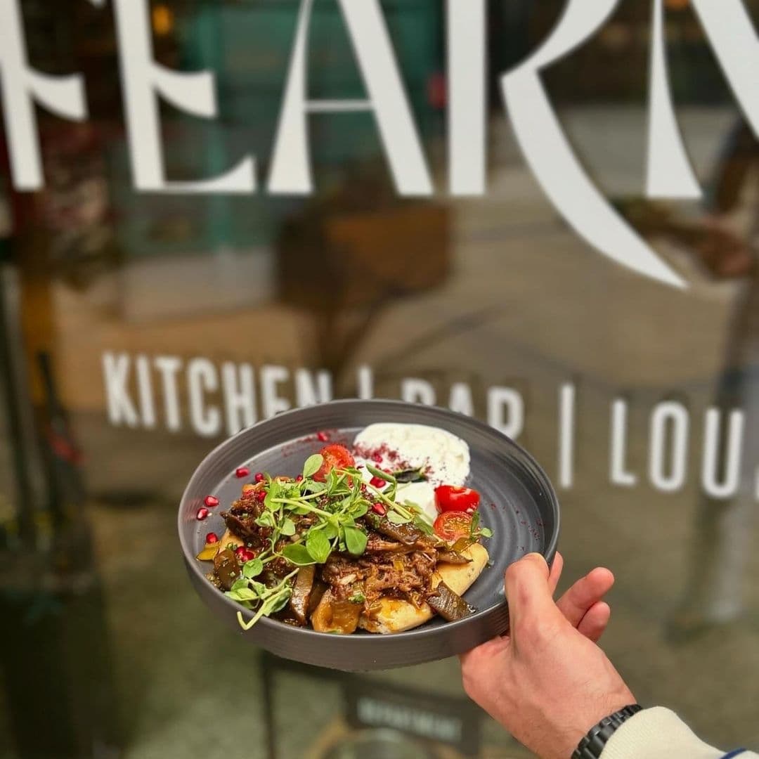 Alt text: "Hand presenting a gourmet dish with garnished meat, fresh greens, and roasted vegetables at Fearns Dock, with the restaurant's sign in the blurred background."
