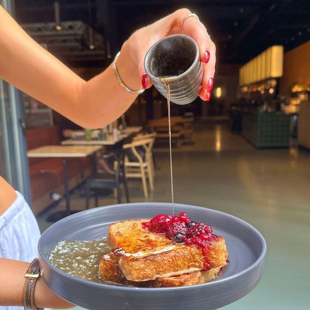 Alt text: "Delicious French toast topped with berries being drizzled with syrup at Fearns Dock, served on a stylish gray plate held by a person with red nail polish."