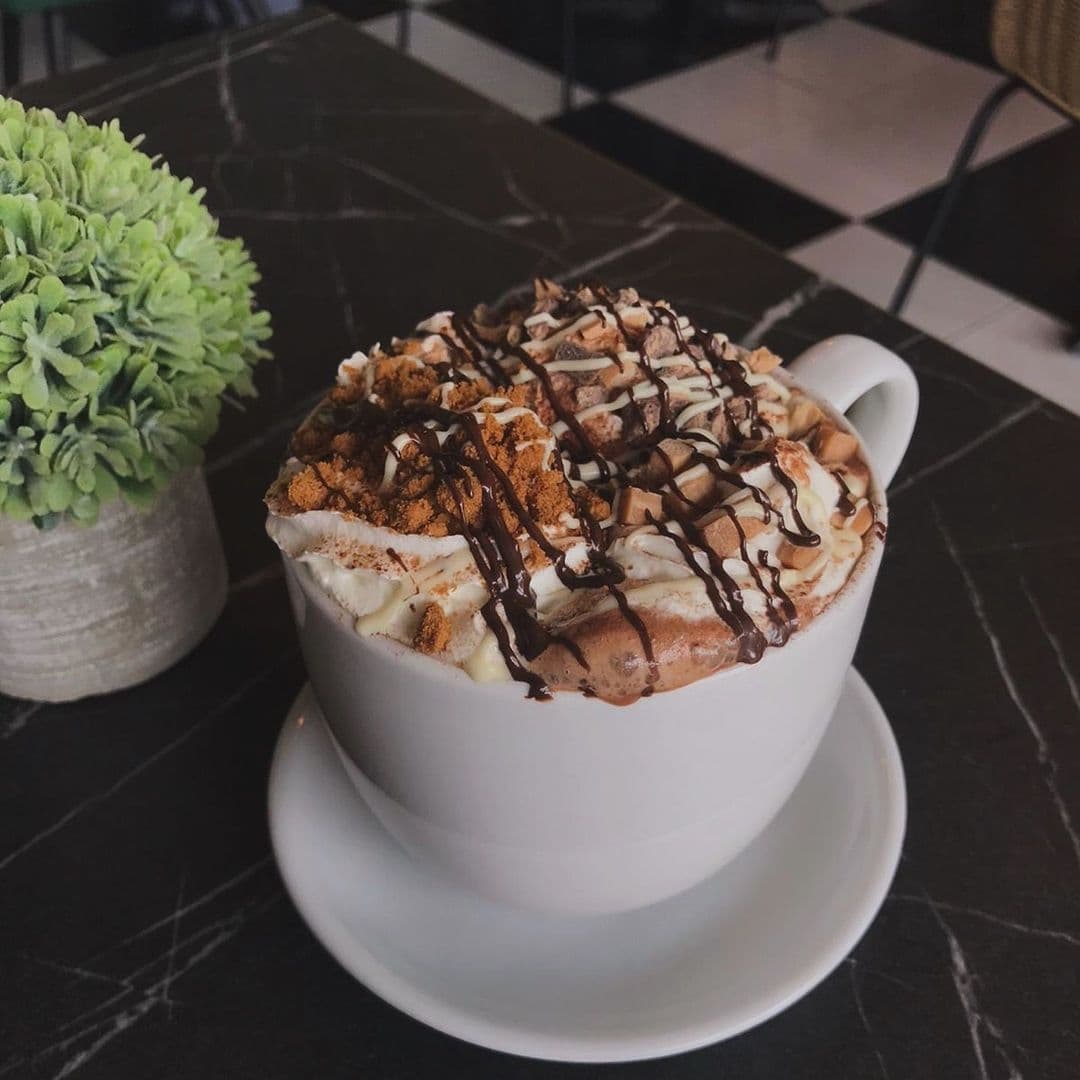 A decadent hot chocolate topped with whipped cream, chocolate syrup, and crumbled cookies, served in a white cup at a cafe in Forista.