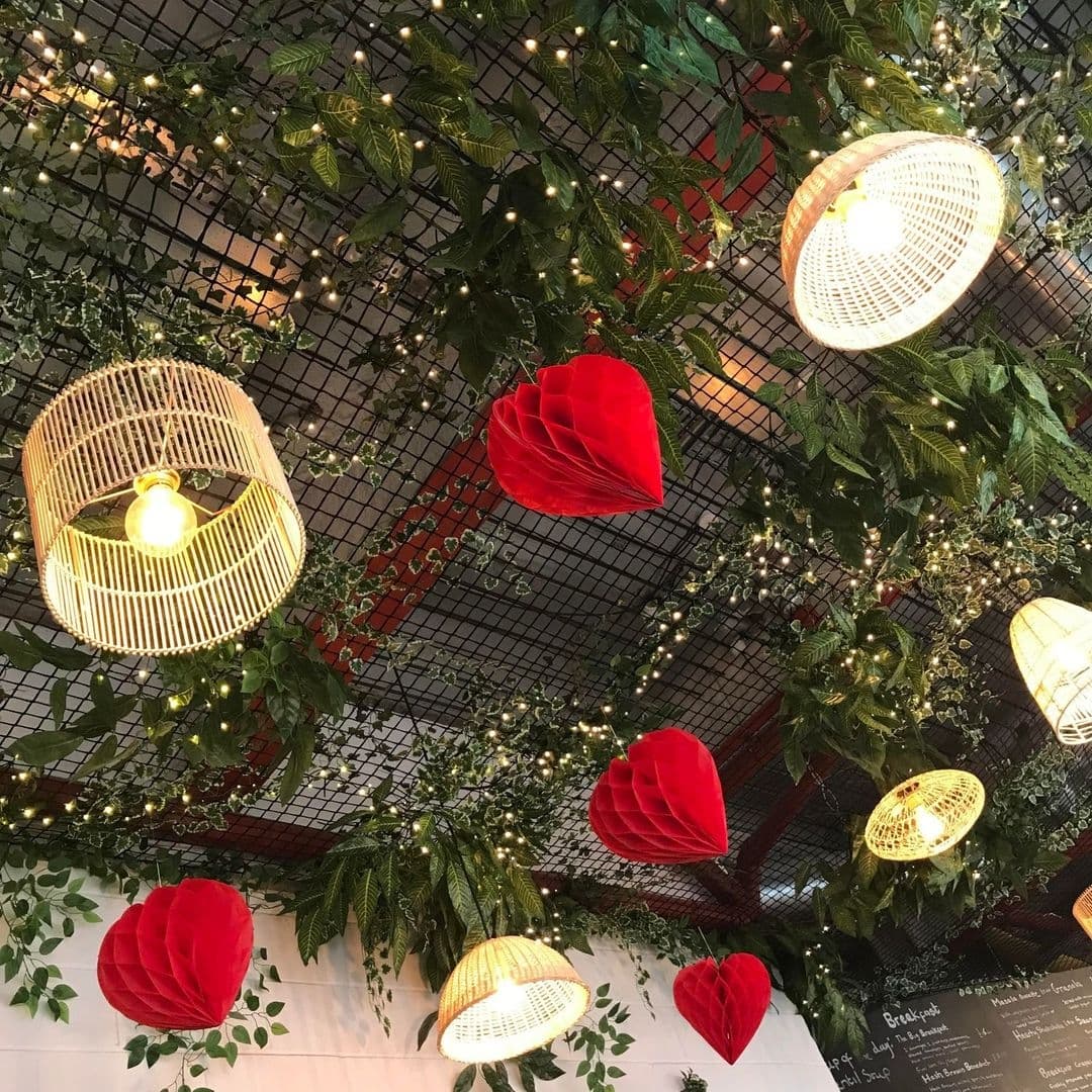 Ceiling decor at Forista featuring white birdcage-style light fixtures and red paper art installations amidst green foliage and twinkling fairy lights.