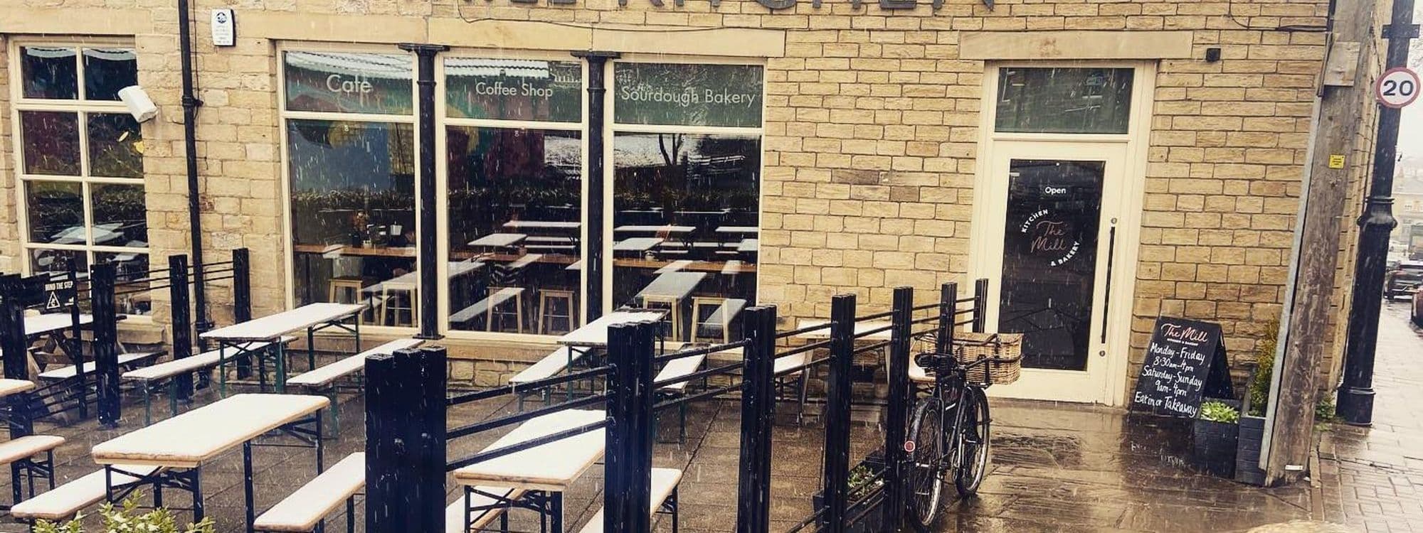 Exterior view of Mill Kitchen & Bakery featuring outdoor seating with snow-covered tables, a bicycle parked by the fence, and visible signage for the café and sourdough bakery.