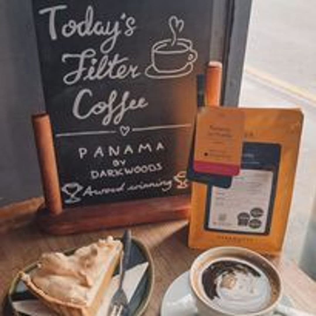 Alt text: "A cozy corner at Pooky’s Deli & Cafe featuring today's filter coffee from Panama, a slice of pie on a plate, and a cup of coffee with latte art on the table, next to a menu stand."