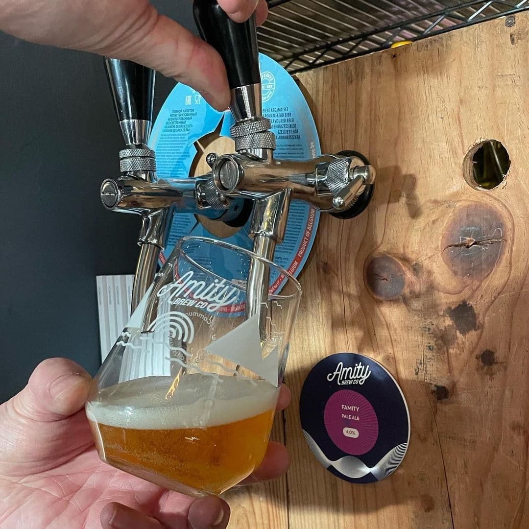Beer being poured from a tap into a glass at Pooky’s Deli & Cafe, with a person's hand on the tap and a coaster for Amity Pale Ale visible.