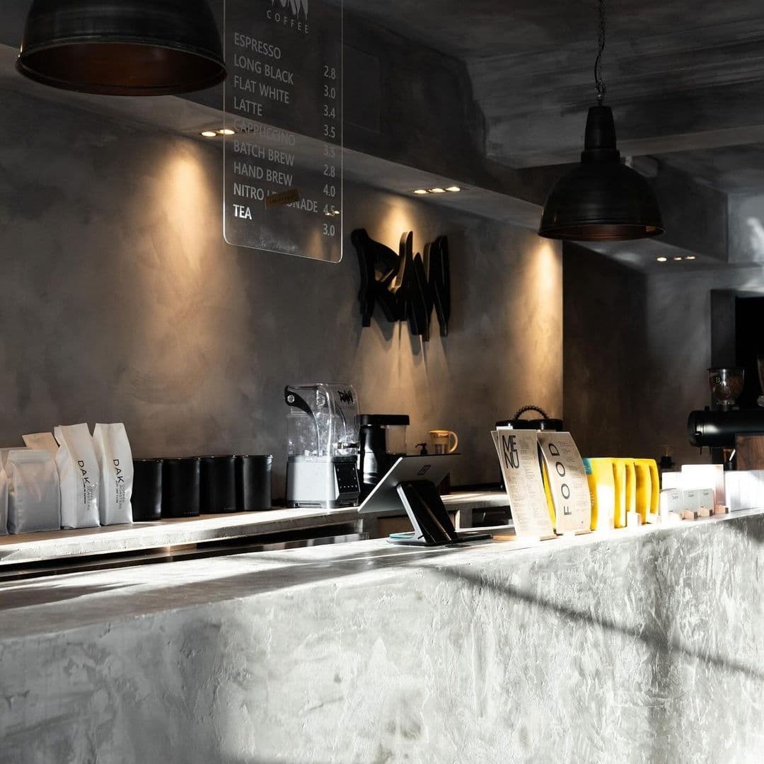 Interior of Raw Coffee shop with a modern, industrial aesthetic featuring a concrete counter, espresso machine, and menu board listing coffee and tea options.