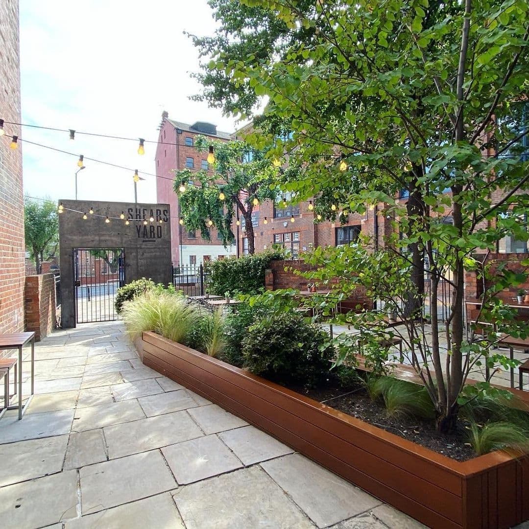 Outdoor patio area at Shears Yard with string lights, green plants, and brick buildings in the background.