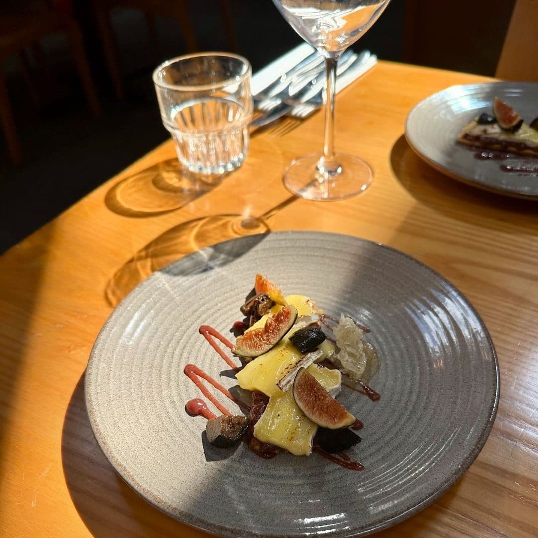 Gourmet dessert plate with figs and cheese at Shears Yard, elegantly presented on a textured ceramic dish, accompanied by a wine glass and water glass on a wooden table with sunlight casting warm shadows.