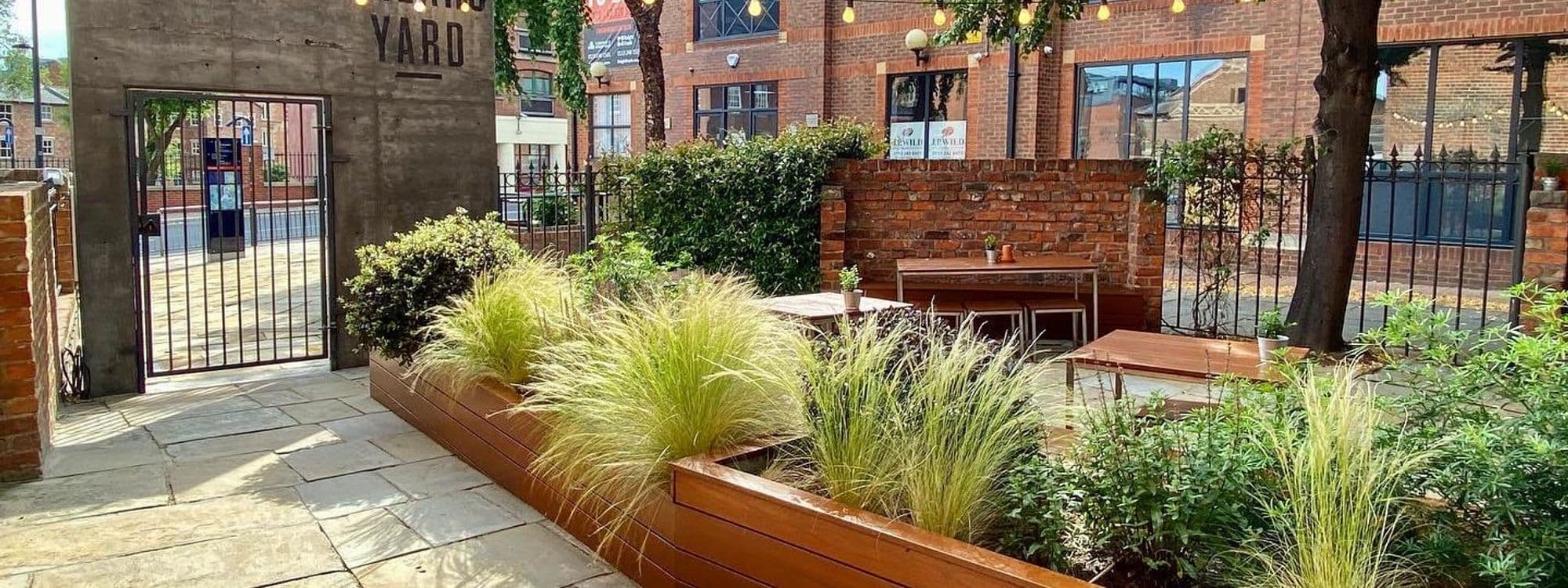 Outdoor seating area at Shears Yard with wooden benches, lush green plants, and a brick wall under a string of lights.