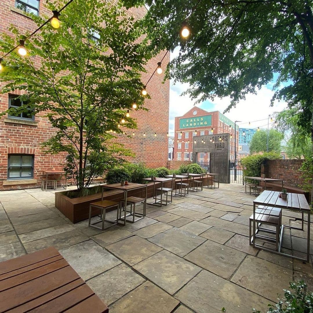 Outdoor seating area at Shears Yard with wooden benches, tables, and string lights, surrounded by brick buildings and greenery.